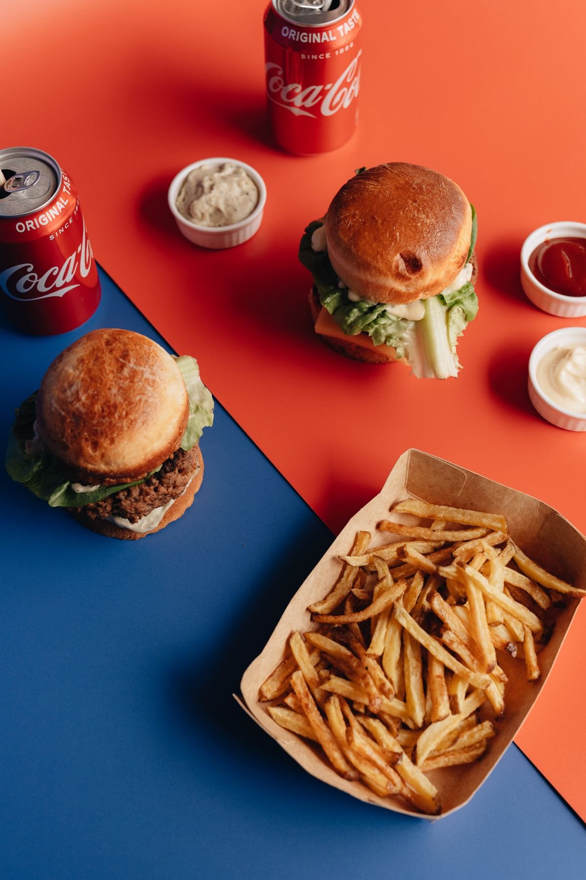Overhead view of burgers, fries and sodas on a bright coloured surface, used as an editorial illustration for “Why burger prices differ by country”